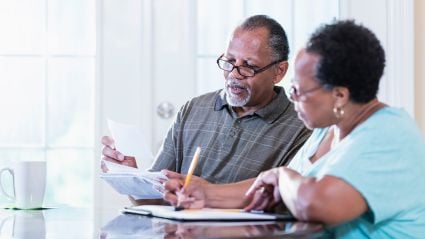 An image of an older couple, a man and woman with brown skin and short brown hair, sitting at a table and reviewing paperwork together. 
