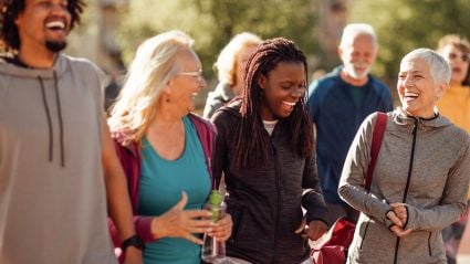 An image of a young man with dark skin, an old woman with white skin, a young woman with dark skin, and another old woman with white skin, standing together and laughing with people and nature in the background. This image leads to an article by the Milken Institute about Scaling and Sustaining Better Health Outcomes Through Prevention.