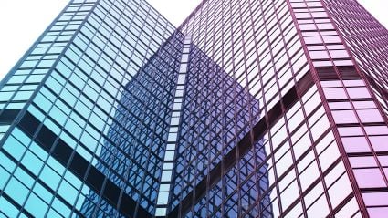 An image of a skyscraper building, upclose, pointing upwards and showing its glass windows with blue-ish and purple-ish hues reflected off of the windows.