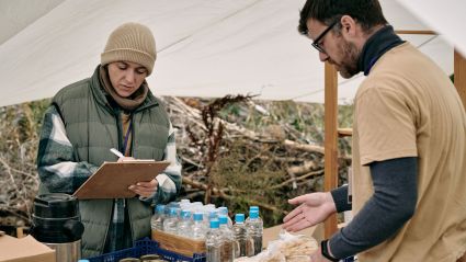 An image of a man and a woman, each with white skin and brown hair, standing under a tent. The woman is checking off a checklist on a clipboard and the man is pointing to a case of water, indicating a disaster relief tent.