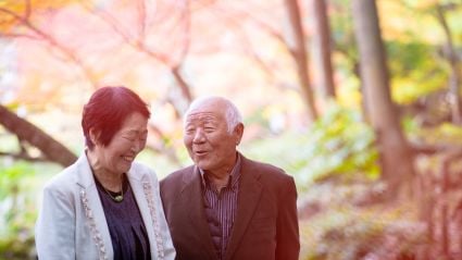 An image of an elderly man and woman who are Asian in appearance with white skin, the woman has short black hair and the man has short white hair. They are walking on a trail underneath trees.