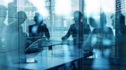 An abstract image with a blue-ish tint of a large window reflection that shows vague figures of business people sitting at a table conducting a meeting.