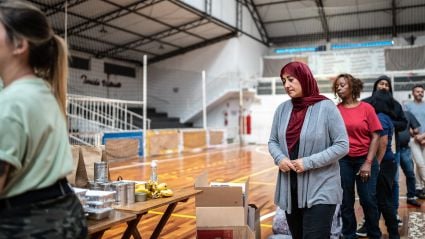 An image of various adults standing in a line in what appears to be a school gymnasium and waiting to collect food from a table, indicating a food bank or food drive.
