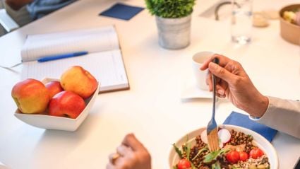 An image of someone's hands eating a bowl of food with various vegetables, and nearby sits a white bowl full of red apples.