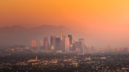 A wide view of Los Angeles with dark smoke billowing from wildfires in the distance, rising behind downtown buildings and palm trees. The sky is streaked with orange and gray, emphasizing the scale of the disaster. The image reflects the urgency and impact of wildfire recovery efforts in the city.