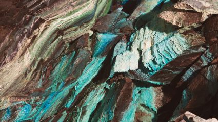 Close-up of jagged rock layers with shimmering turquoise, teal, and brown mineral streaks creating a textured, iridescent surface.