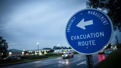 Blue emergency management sign showing an evacuation route with a left-pointing arrow, positioned along a wet roadway at dusk with cars and buildings in the background.