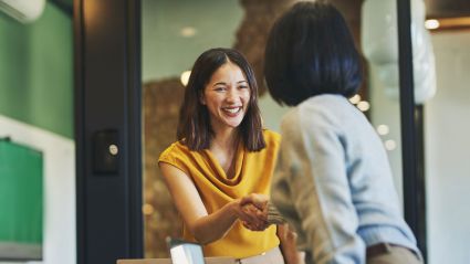 Two people in a professional setting smiling and shaking hands, symbolizing agreement or partnership.