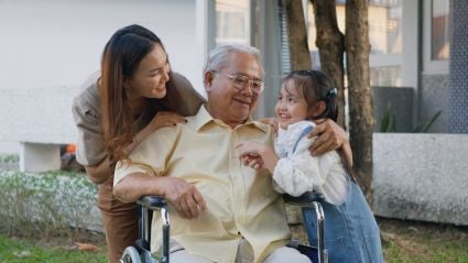 An elderly man in a wheelchair smiles warmly as he is embraced by a young woman and a little girl outdoors, sharing a joyful family moment together.