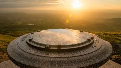 Stone compass viewpoint on a hilltop at sunset, with golden sunlight casting a warm glow over the engraved circular map and the rolling countryside landscape in the distance.