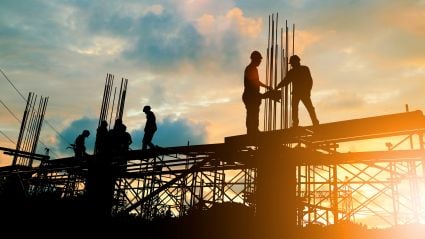 Silhouetted construction workers on elevated scaffolding at sunset, adjusting rebar and walking across metal framework against an orange-blue sky.