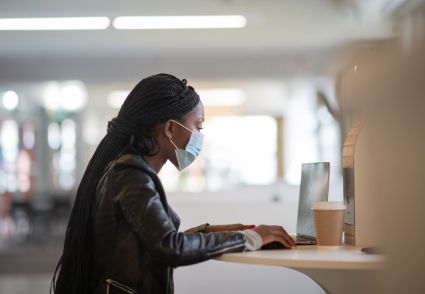 woman wearing mask, working at laptop with coffee on table
