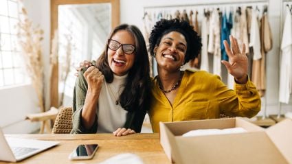 Two women smiling and waving while seated at a table in a studio, with clothing racks visible in the background.
