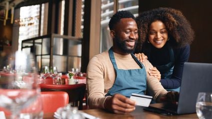 Two small business owners smiling while using a laptop and credit card in their restaurant, representing financial growth and community lending success.
