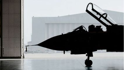A dramatic silhouette of a fighter jet parked inside a hangar, with the cockpit canopy open and a misty airfield visible through the large hangar doors.