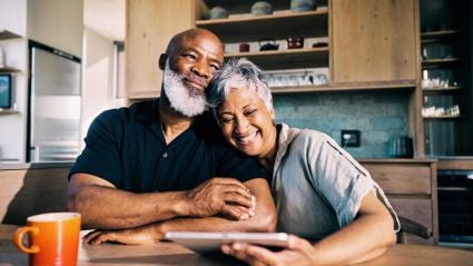 A smiling older Black man with a white beard and a woman with short grey hair share an affectionate moment in a kitchen.