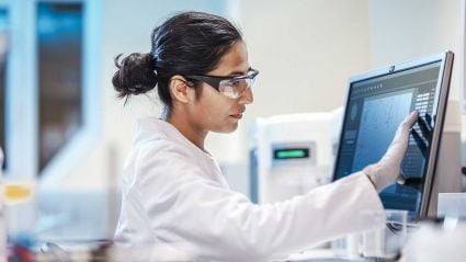 A female scientist in a white lab coat and safety goggles focuses intently while interacting with a touch-screen monitor in a modern laboratory setting.