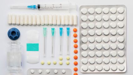 A high-angle, organized flat lay of pharmaceutical supplies on a white background, including a syringe, a large blister pack of white pills, a row of capsules, sterile needles, and loose orange tablets.