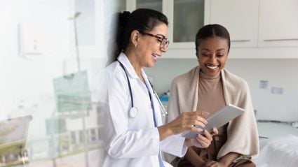 A doctor in a white coat and stethoscope smiles while showing a tablet to a patient seated on an exam table in a medical office. Both women are smiling and engaged with the screen.