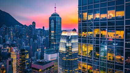Modern glass office towers in Hong Kong at dusk with illuminated interiors and surrounding urban skyline against a colorful sunset.