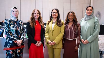 Five professionally dressed women from diverse backgrounds stand together smiling in a conference room.