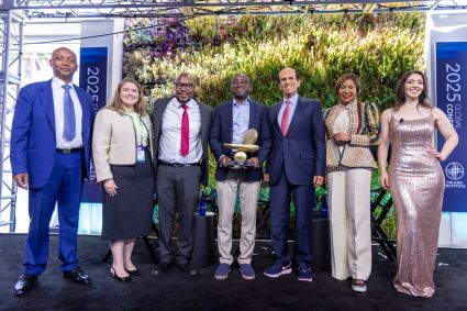 Seven people pose on stage at the 2025 Milken Institute Global Conference, with one person holding a gold award trophy.