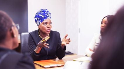 A woman in a blue patterned head wrap speaks animatedly during a business meeting with colleagues.