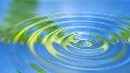 Close-up of concentric water ripples reflecting blue sky and green foliage.