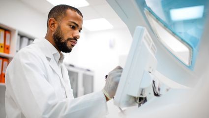 A scientist in a white lab coat and gloves operating medical or laboratory equipment in a bright clinical setting.