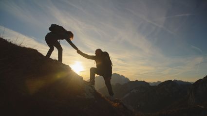 Two hikers silhouetted against a sunset, one reaching down to help the other climb a rocky mountain slope.