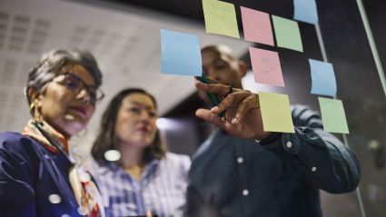 Three colleagues collaborating at a glass wall covered in colorful sticky notes during a brainstorming session.