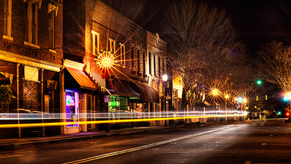 Historic downtown street at night with illuminated storefronts and light trails from passing traffic
