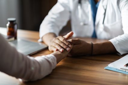 Doctor in white coat with stethoscope holding patient's hands across wooden desk