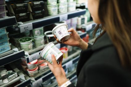 Person in dairy aisle of grocery store comparing two containers of what appears to be yogurt