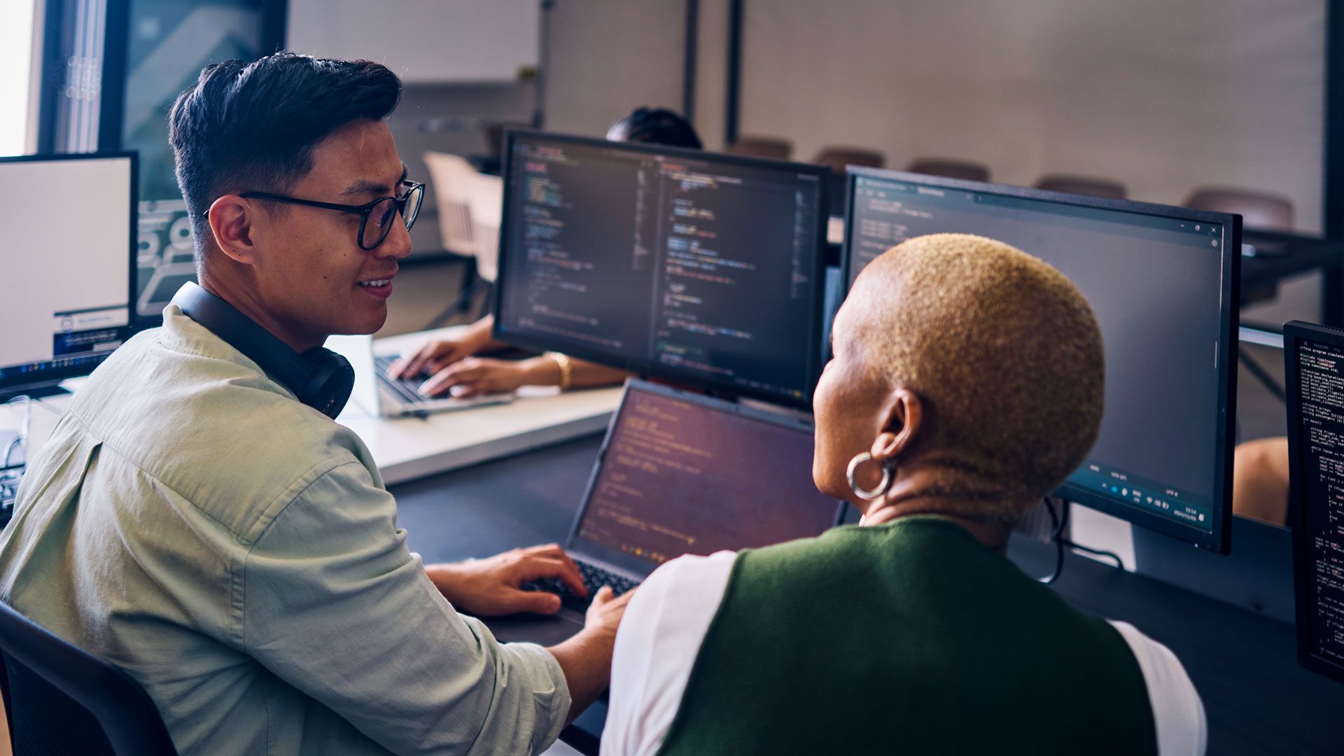 Two colleagues collaborate at a desk, reviewing code on multiple computer monitors.