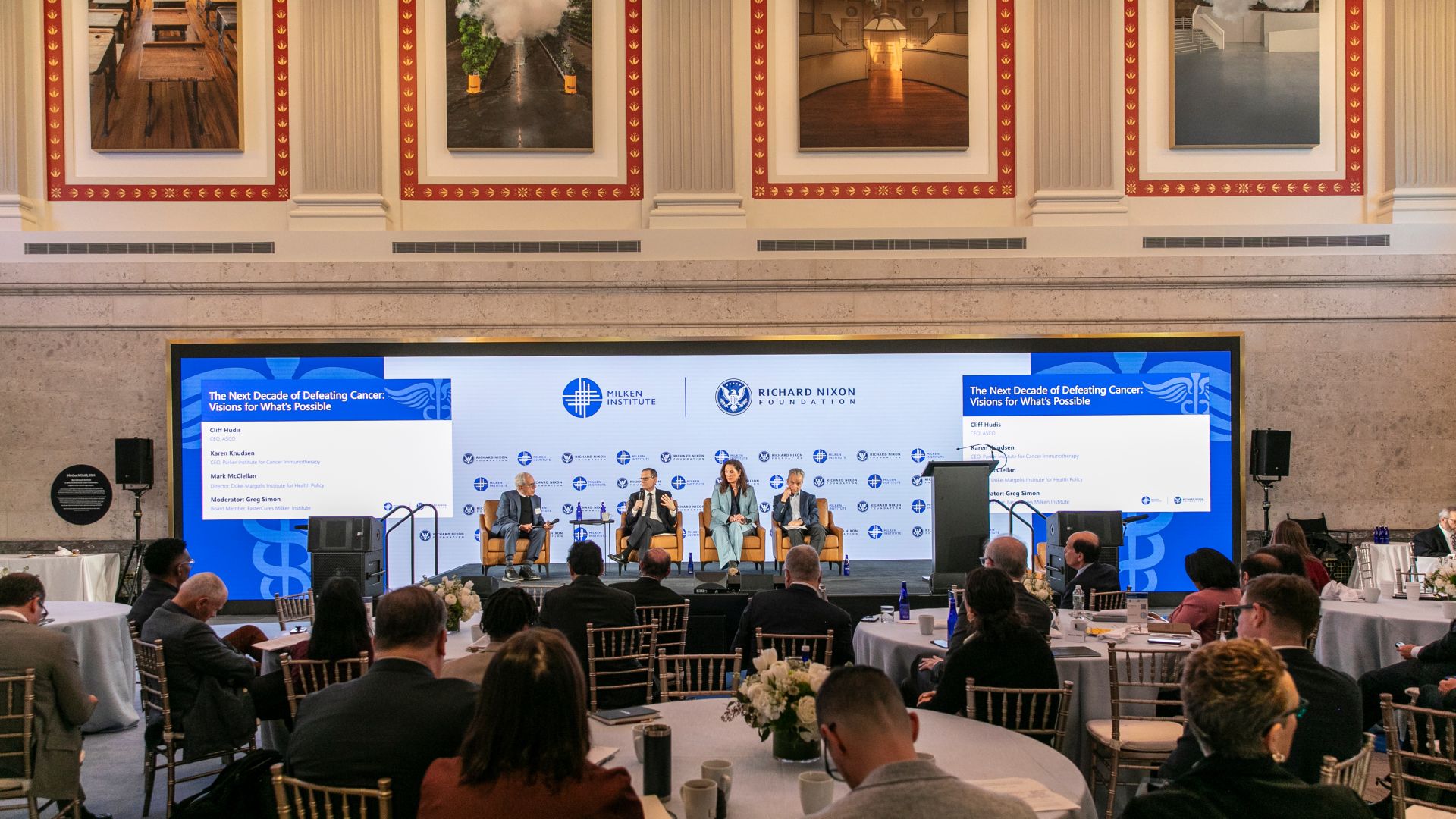 Panel discussion on stage with large blue screens in elegant ballroom with chandeliers