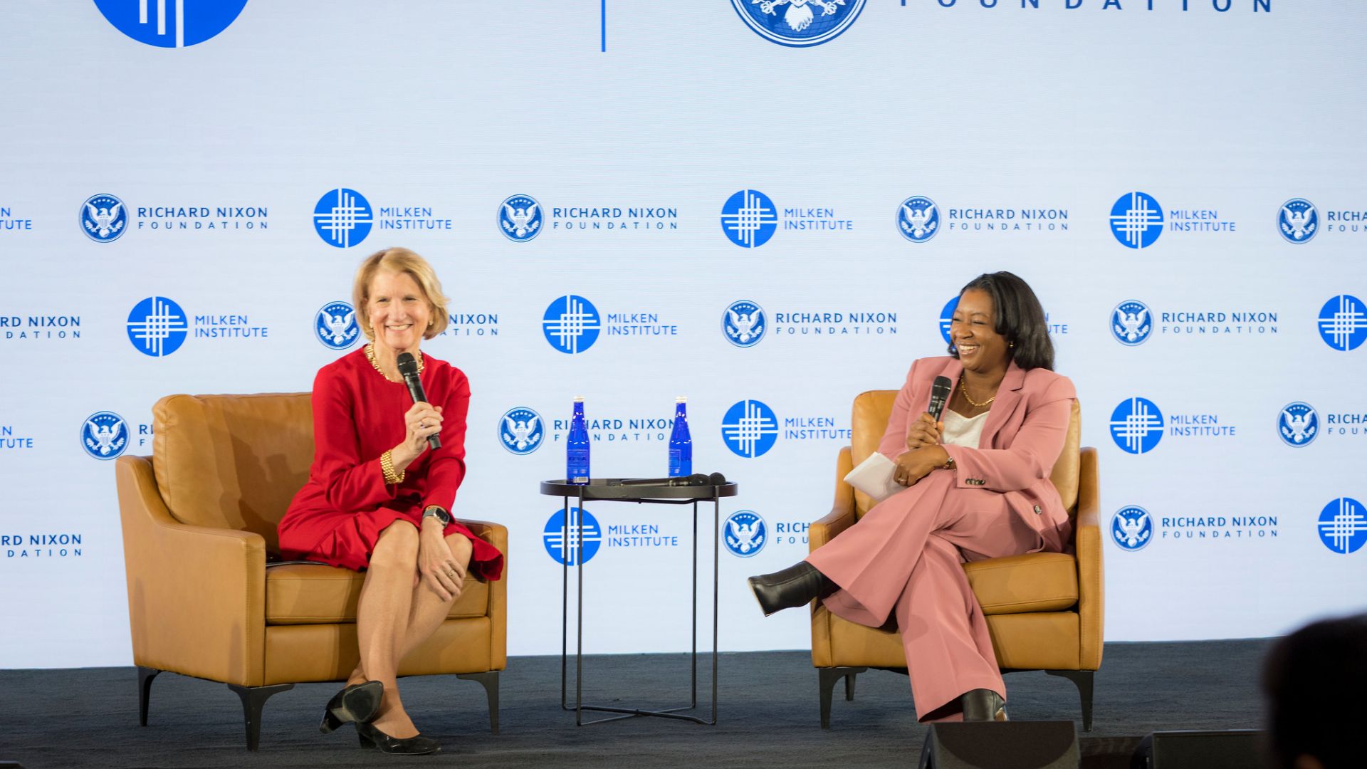 Two women in business attire seated in armchairs on stage during a panel discussion with branded backdrop