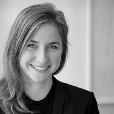 Smiling light-skinned, female-presenting individual with long length blonde hair wearing a black shirt on a grey backdrop.