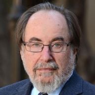 A light-skinned male-presenting person with short, dark hair and a beard wearing a suit against a blurred brown background