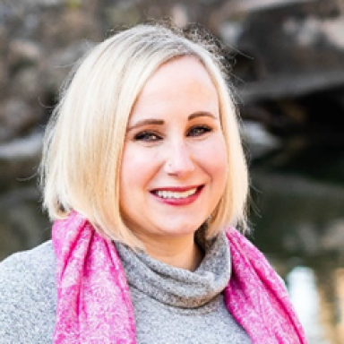 A smiling light-skinned female-presenting person with light hair wearing a gray sweater and pink scarf against a blurred background