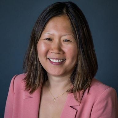 A smiling female-presenting person with dark hair in a pink suit jacket against a dark background. 