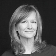 A smiling light-skinned female-presenting person with shoulder-length hair in a black and white picture with a black background