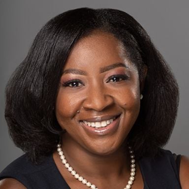 A smiling dark-skinned female-presenting individual with black hair wearing a black shirt and a pearl necklace against a dark gray gradient background