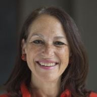 A smiling light-skinned female-presenting person with long, dark hair wearing a red shirt against a dark, blurred background