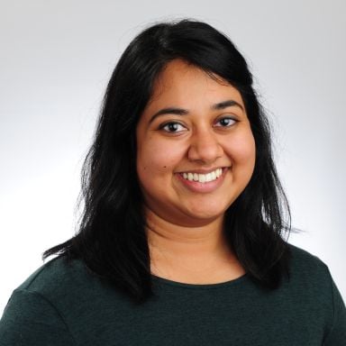 Smiling female-presenting person of color with dark hair wearing a dark green shirt against a light gray background