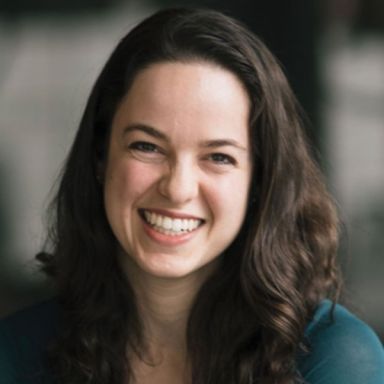 A smiling light-skinned, female-presenting individual with dark brown hair wearing a dark green shirt in front of a blurred background