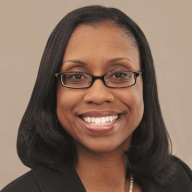 A smiling dark-skinned female-presenting person with black hair wearing a dark suit jacket and glasses against a light brown background