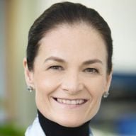 A smiling light-skinned female-presenting person withdark hair wearing a black turtleneck shirt against a blurred background