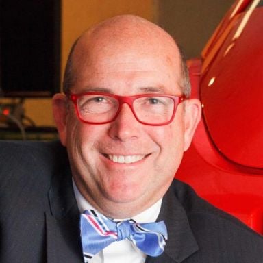 A male-presenting person wearing red eye glasses and a bow tie against a warm background.