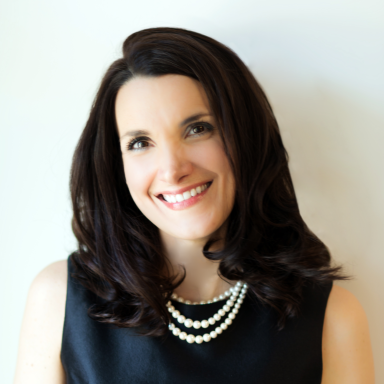 A smiling light-skinned female-presenting person with dark brown hair wearing a black shirt with a pearl necklace against a light gray background